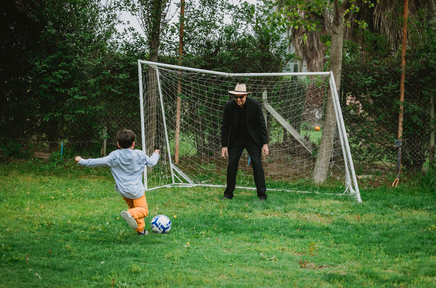 A child kicks a soccer ball toward an adult goalkeeper in a backyard goal on a green lawn.