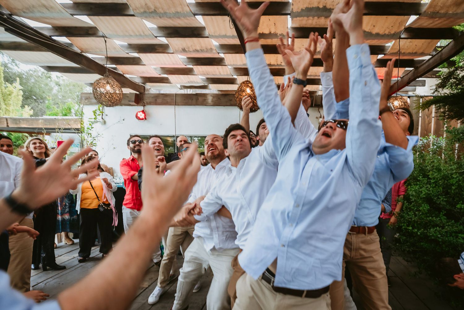 Group of men in blue shirts reaching up to catch bouquet at outdoor wedding reception.