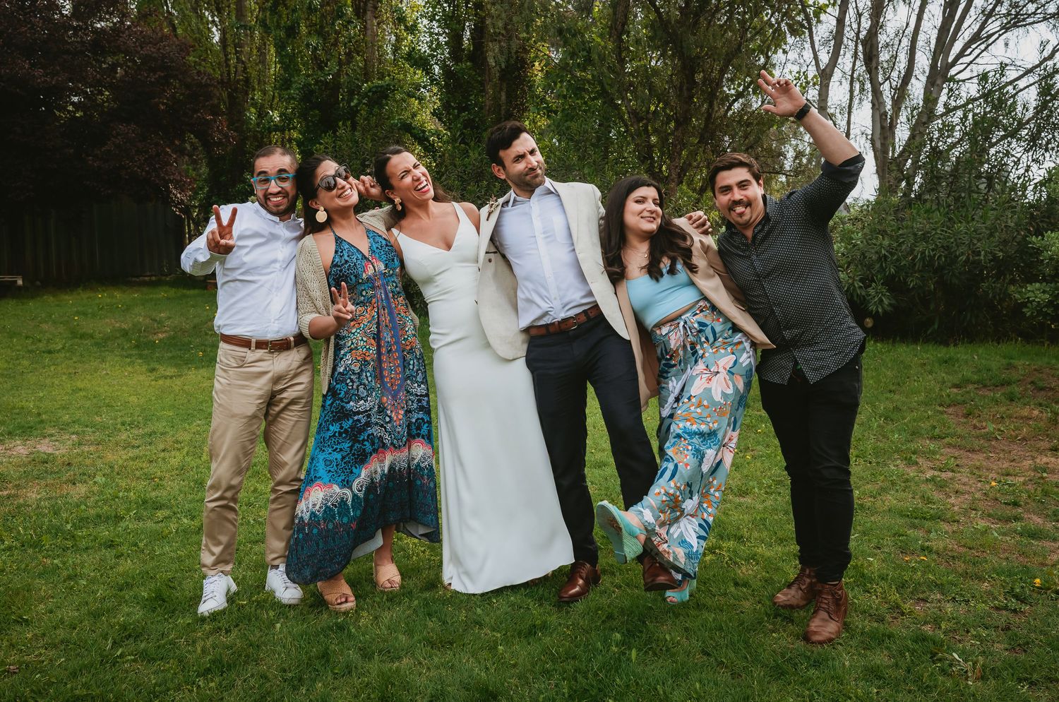 Wedding party of six posing joyfully on green lawn, bride in white gown surrounded by guests in colorful attire.