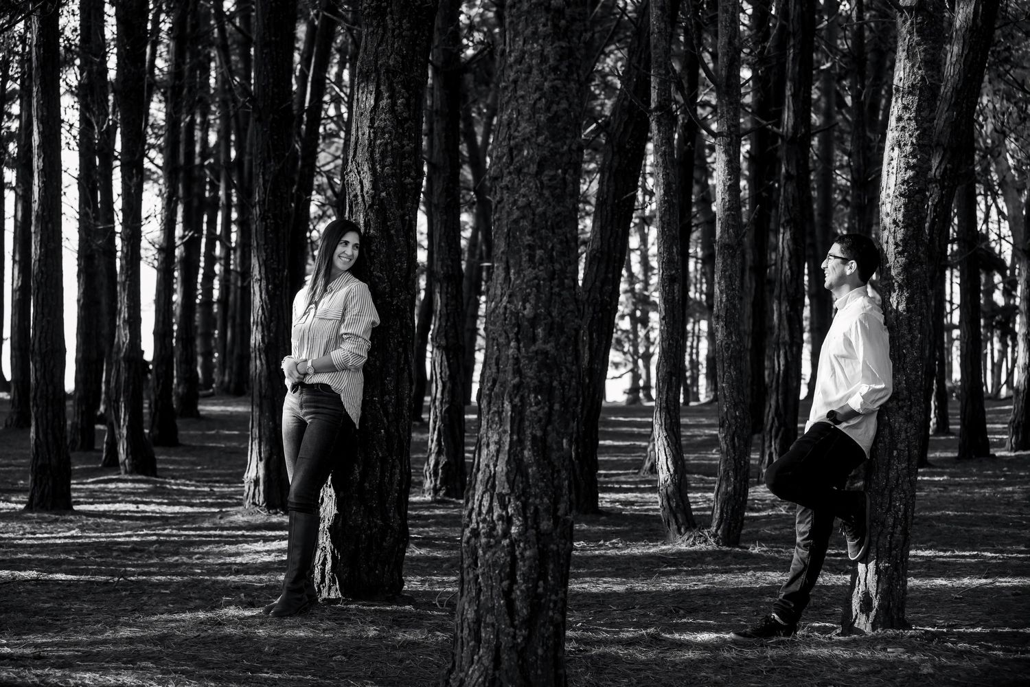 Black and white photo of a couple leaning against separate pine trees, gazing at each other in a forest.