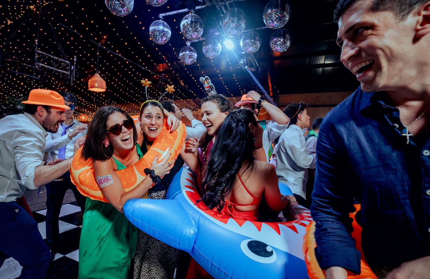 Guests dancing with inflatable shark and pool floats under disco balls at a wedding reception.
