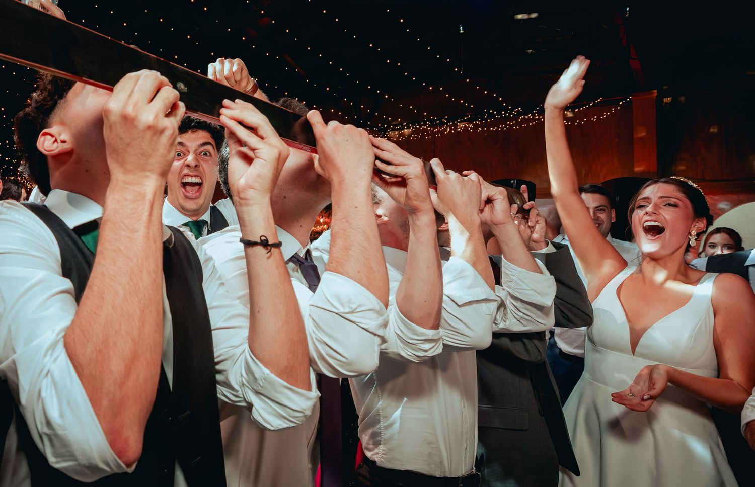 Bride and groom celebrate with wedding guests, raising hands and cheering together at reception.
