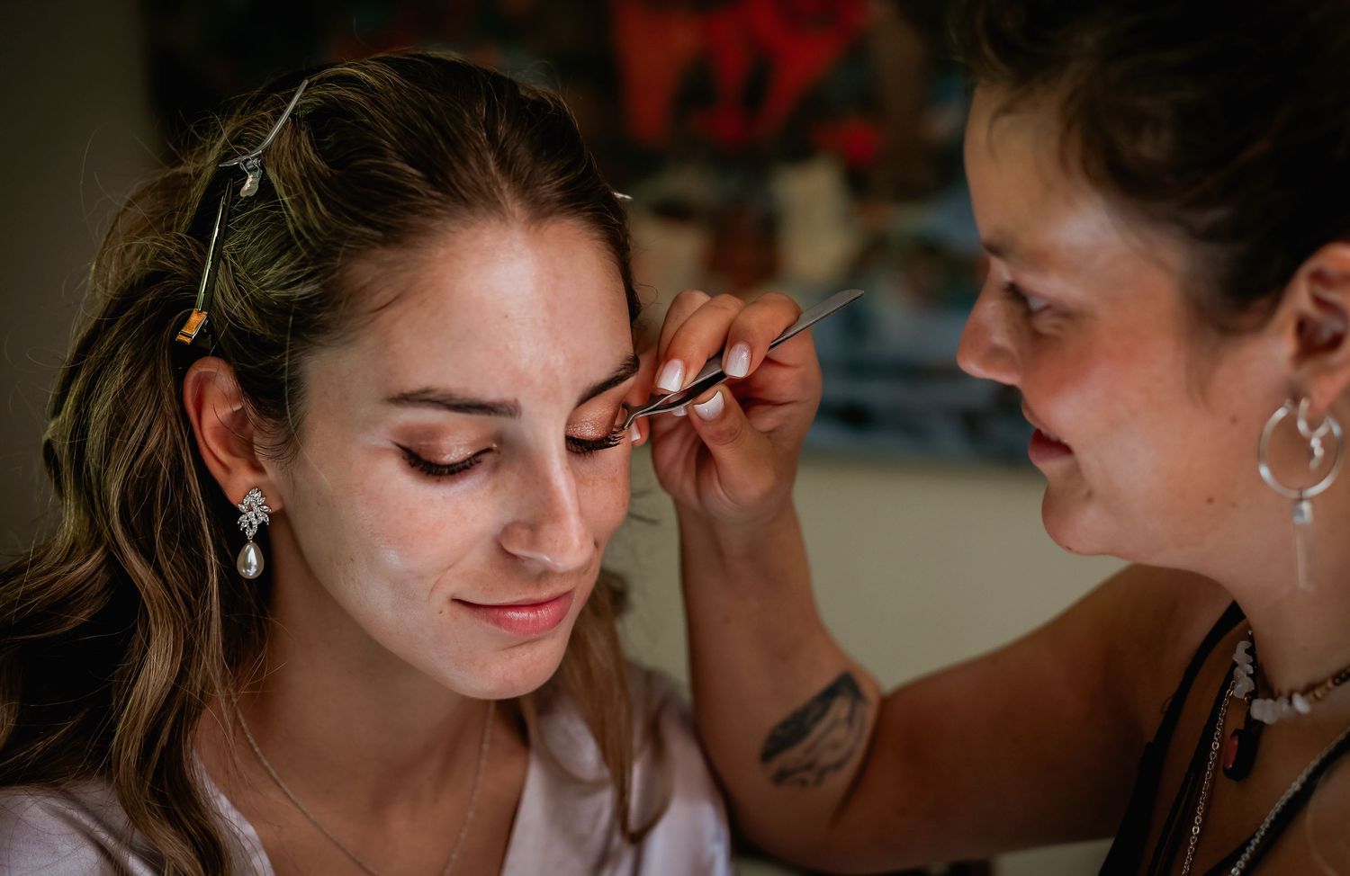 Close-up of makeup artist carefully applying false eyelashes to bride wearing pearl earrings before wedding.