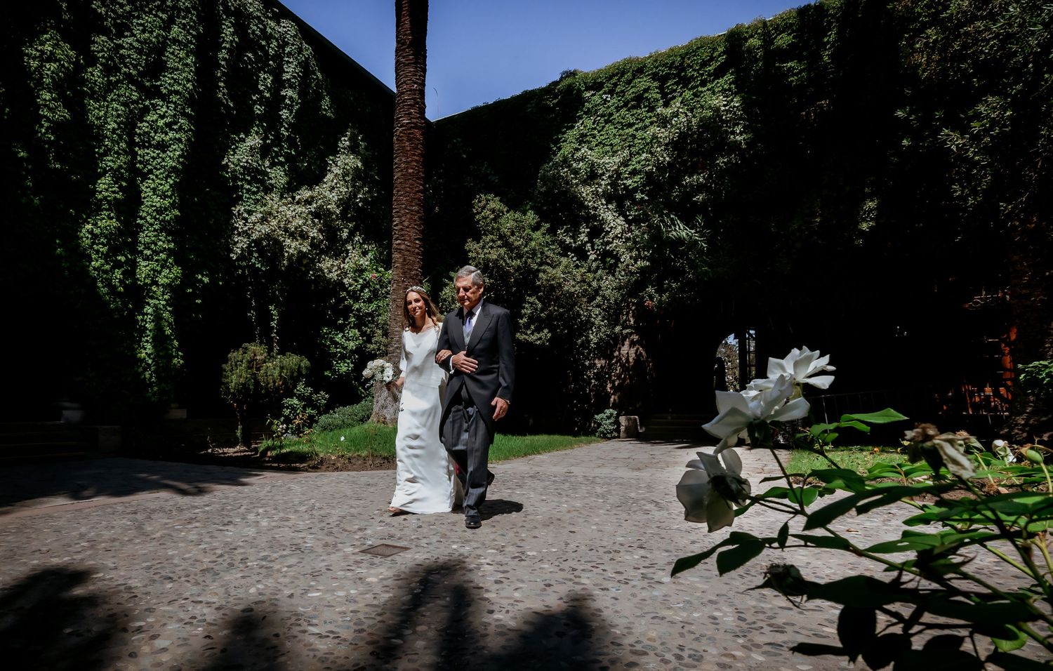 Bride and escort walking cobblestone garden path lined with ivy-covered walls and white roses.