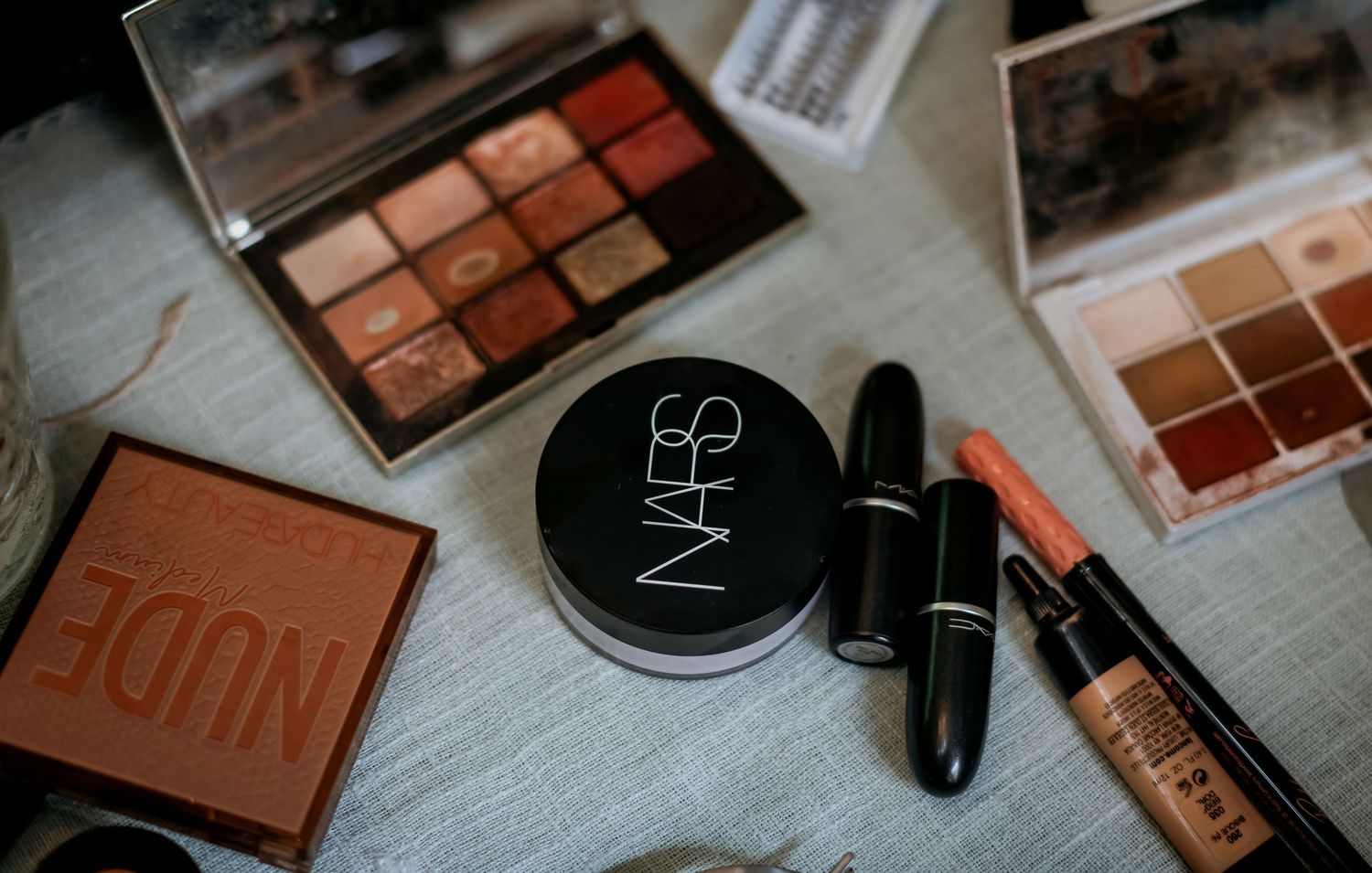 Flat lay of NARS setting powder, nude eyeshadow palette, lipstick, and makeup products on light surface.