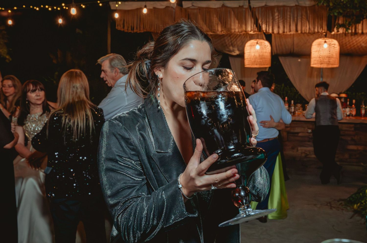 Woman drinks from large glass vessel at outdoor evening wedding reception party.