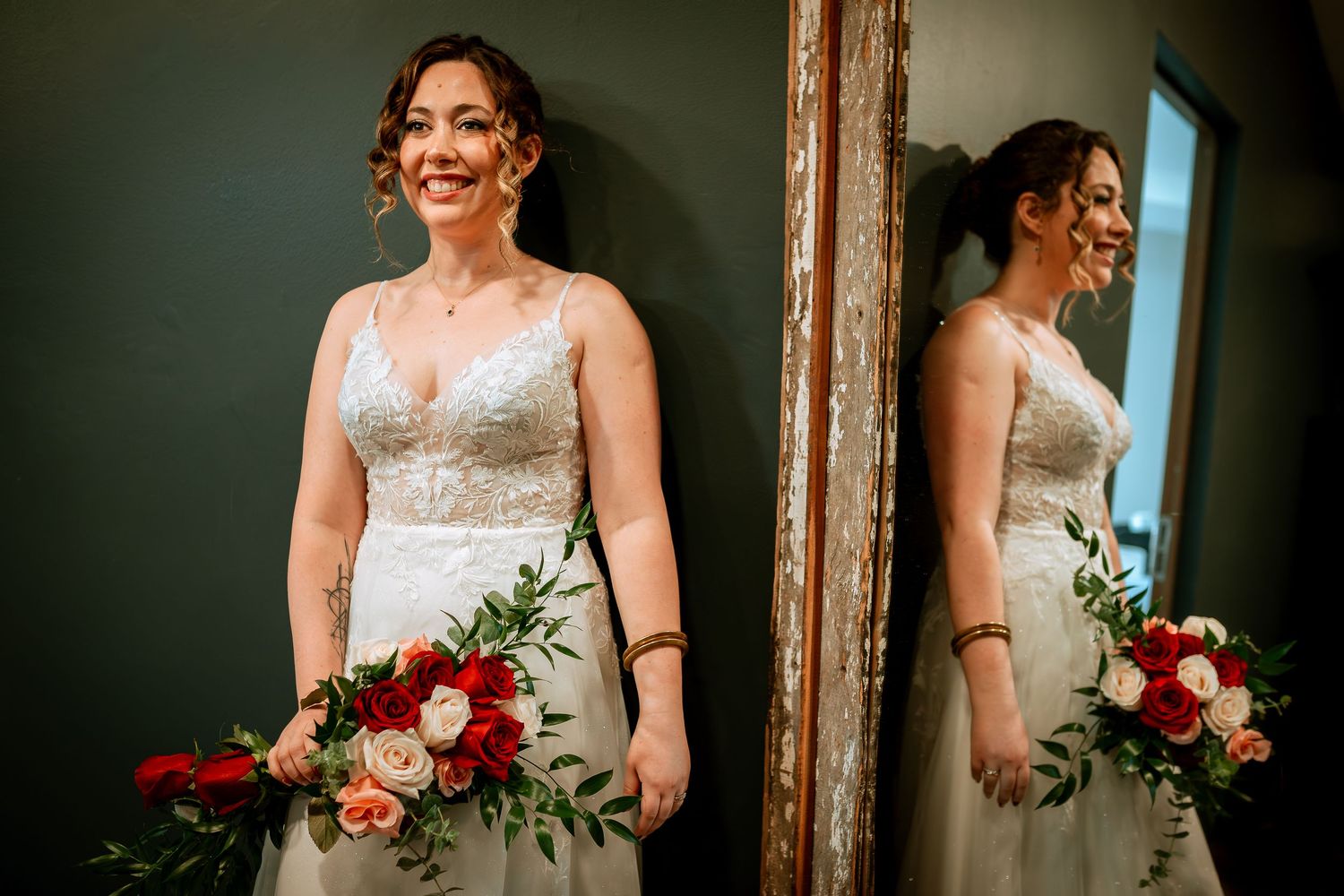Beautiful bride in lace gown holds floral bouquet with her reflection visible in ornate mirror.