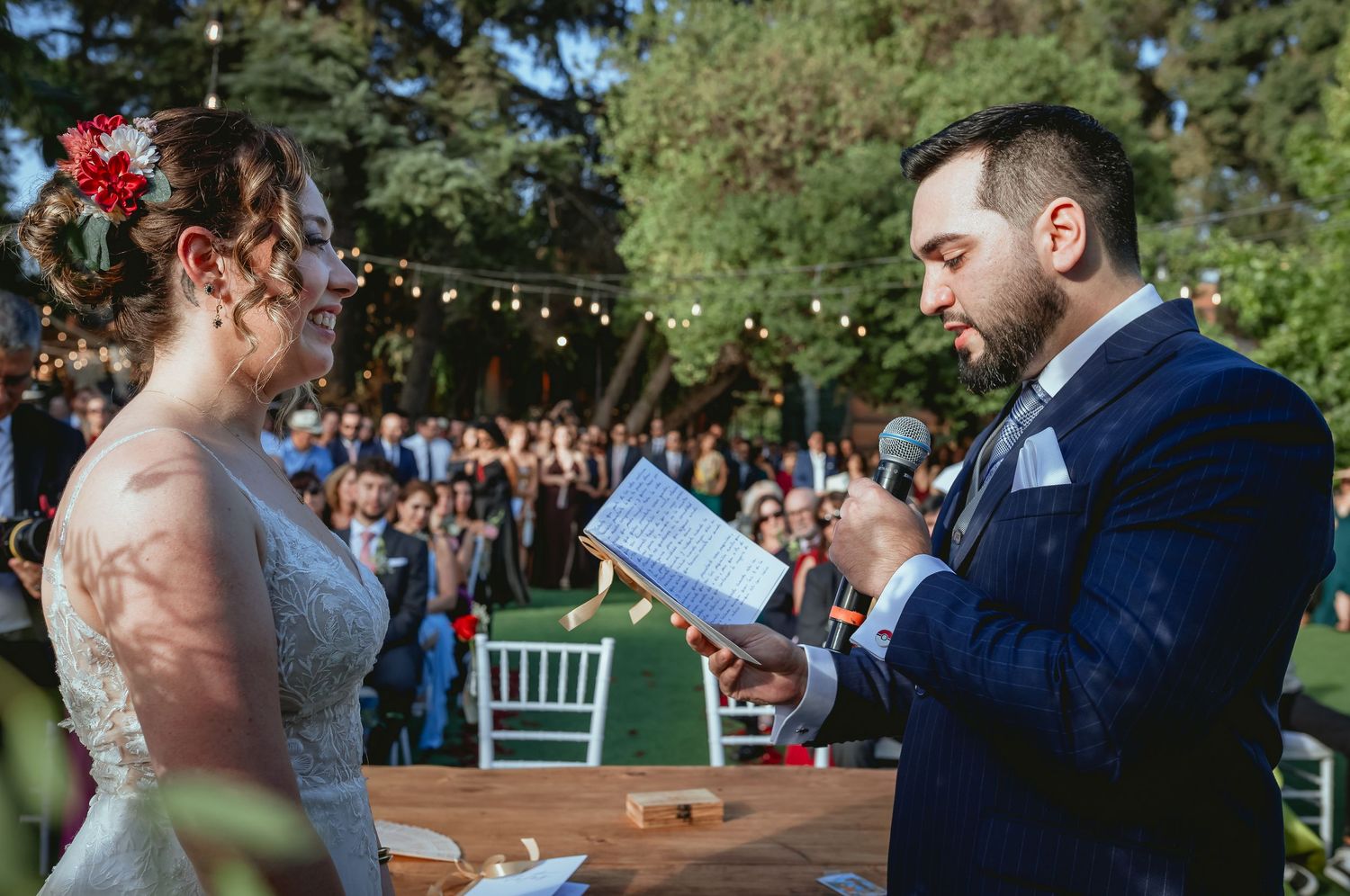 Groom reads wedding vows to bride at outdoor ceremony with guests seated in background.