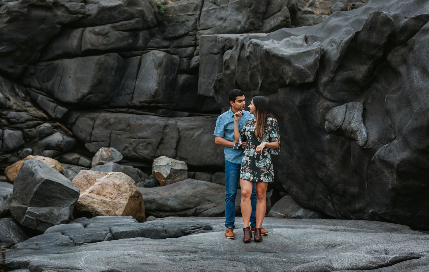 Romantic couple embracing among dark grey coastal boulders in a scenic outdoor setting.