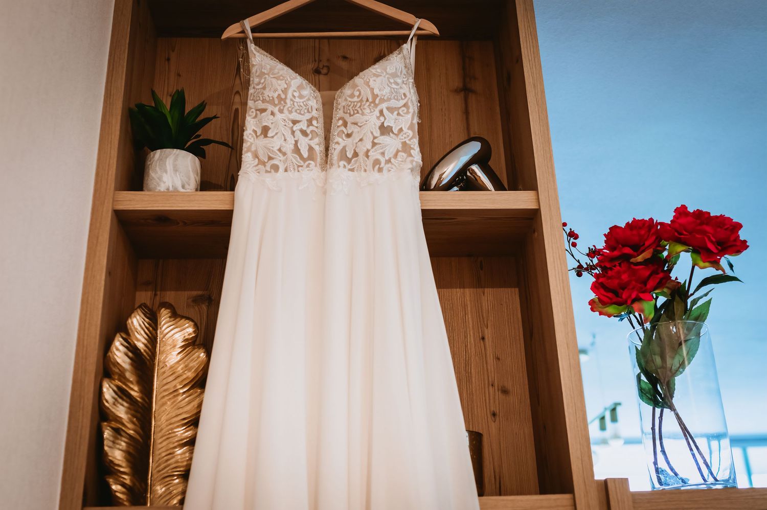 Elegant lace wedding dress hanging on wooden shelf beside red roses and gold decor.