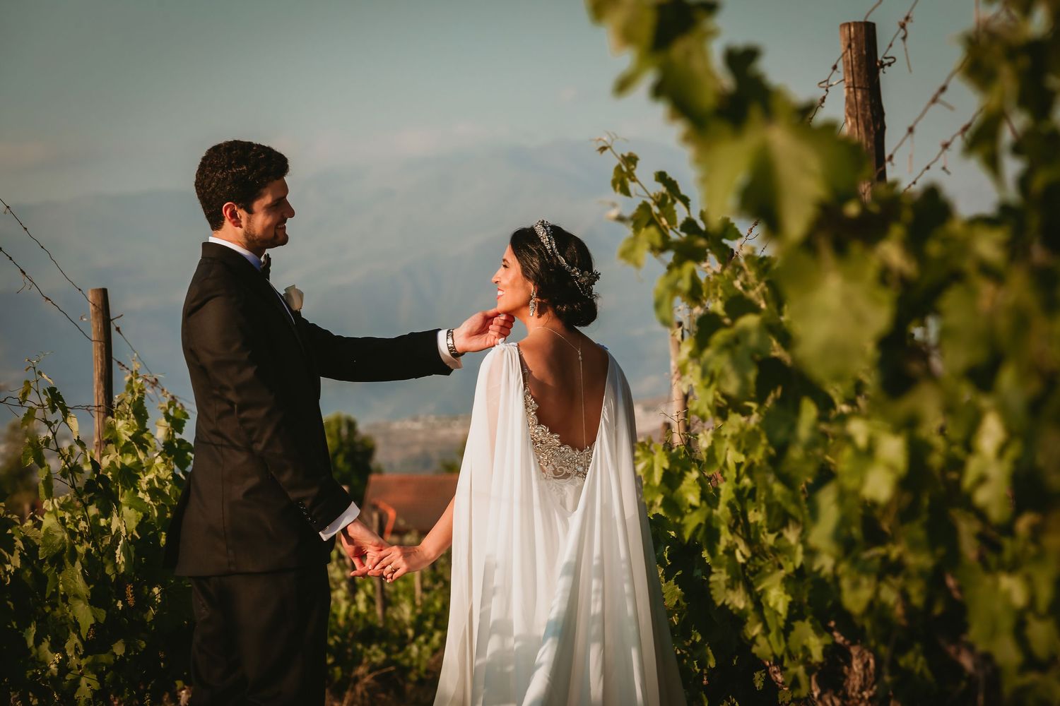 Groom tenderly touches bride's neck as they share an intimate moment in a sunlit vineyard setting.