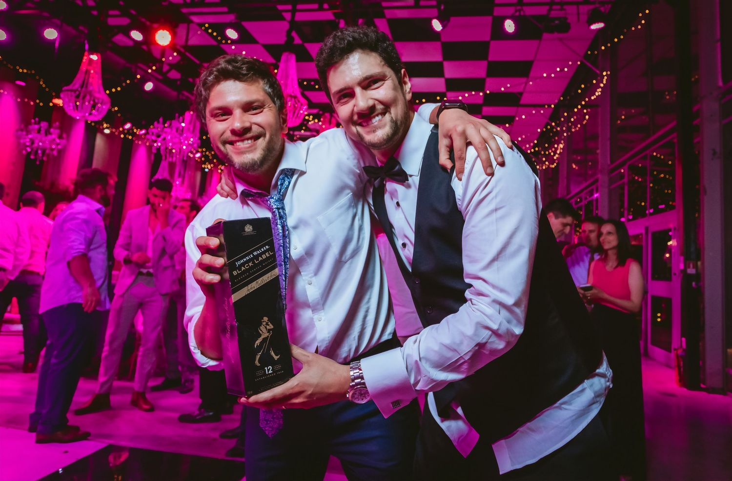 Two men smiling and posing with a Johnnie Walker whiskey bottle at a wedding reception.