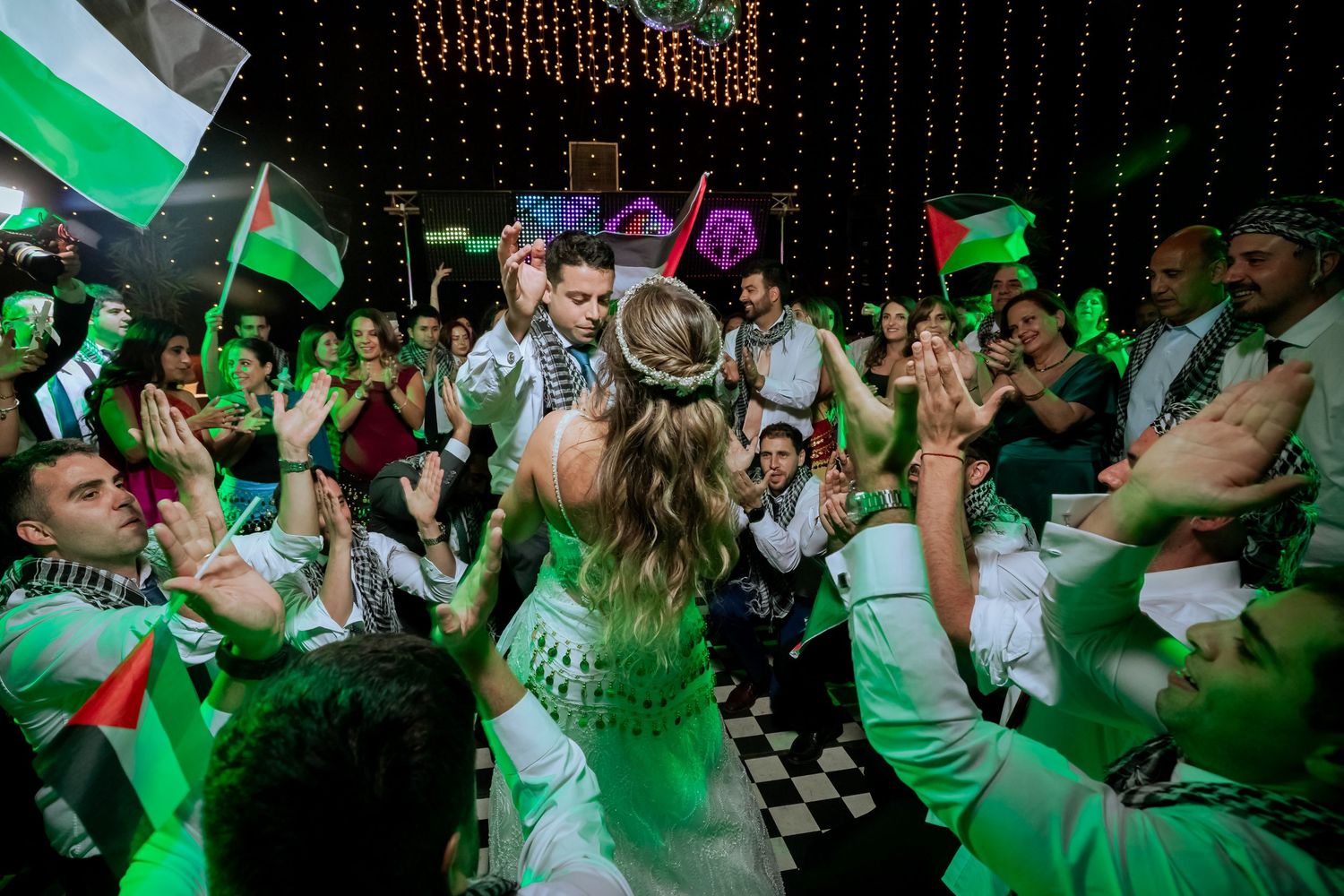 Bride and groom share a kiss on the dance floor surrounded by guests waving Palestinian flags at their wedding.