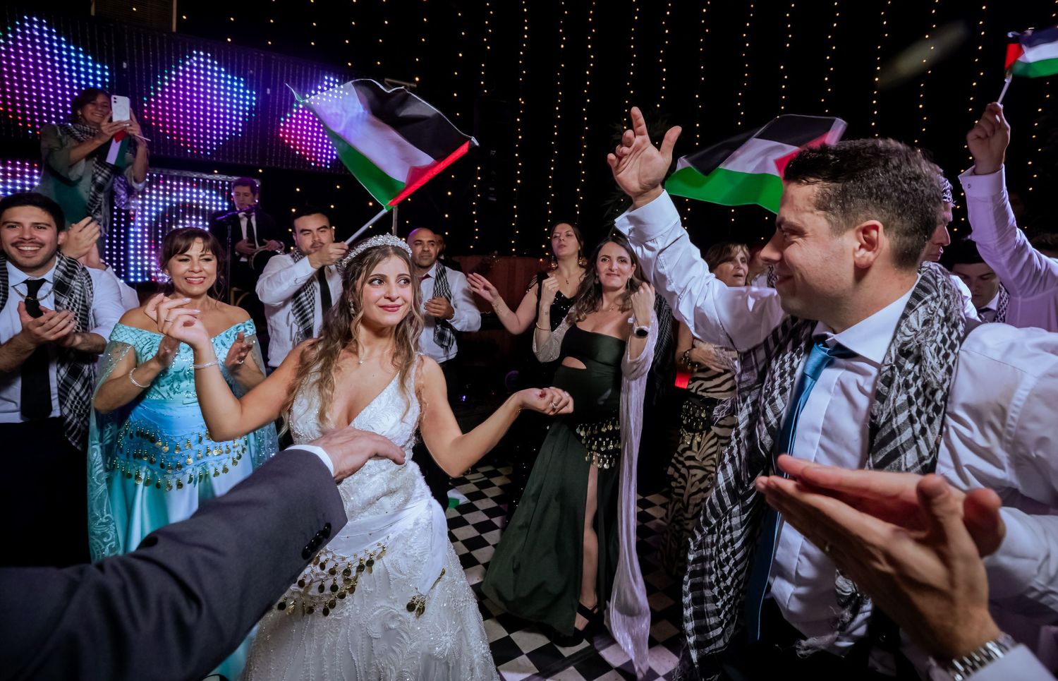 Bride and groom dance joyfully with guests waving Palestinian flags on a checkered dance floor at their wedding.