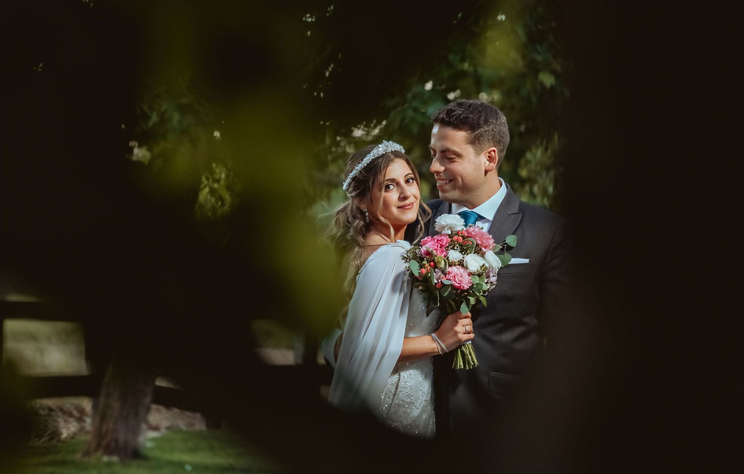 Romantic bride and groom portrait framed by lush green foliage, bride holding a colorful bouquet.