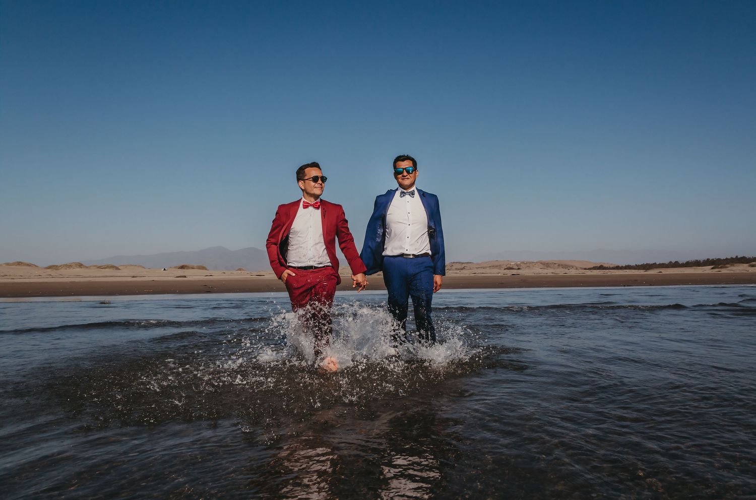 Two grooms in red and blue suits splash through shallow water in a vast sandy landscape.