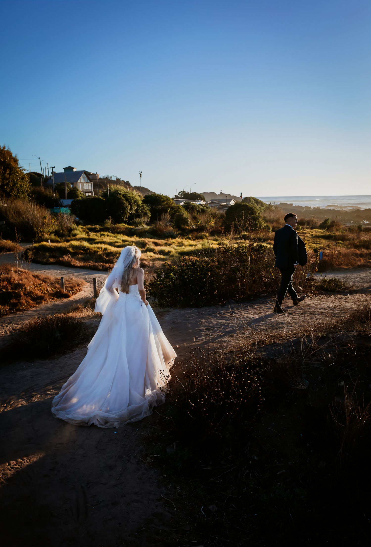 Bride in flowing white gown and groom walk along scenic coastal path toward ocean at golden hour.