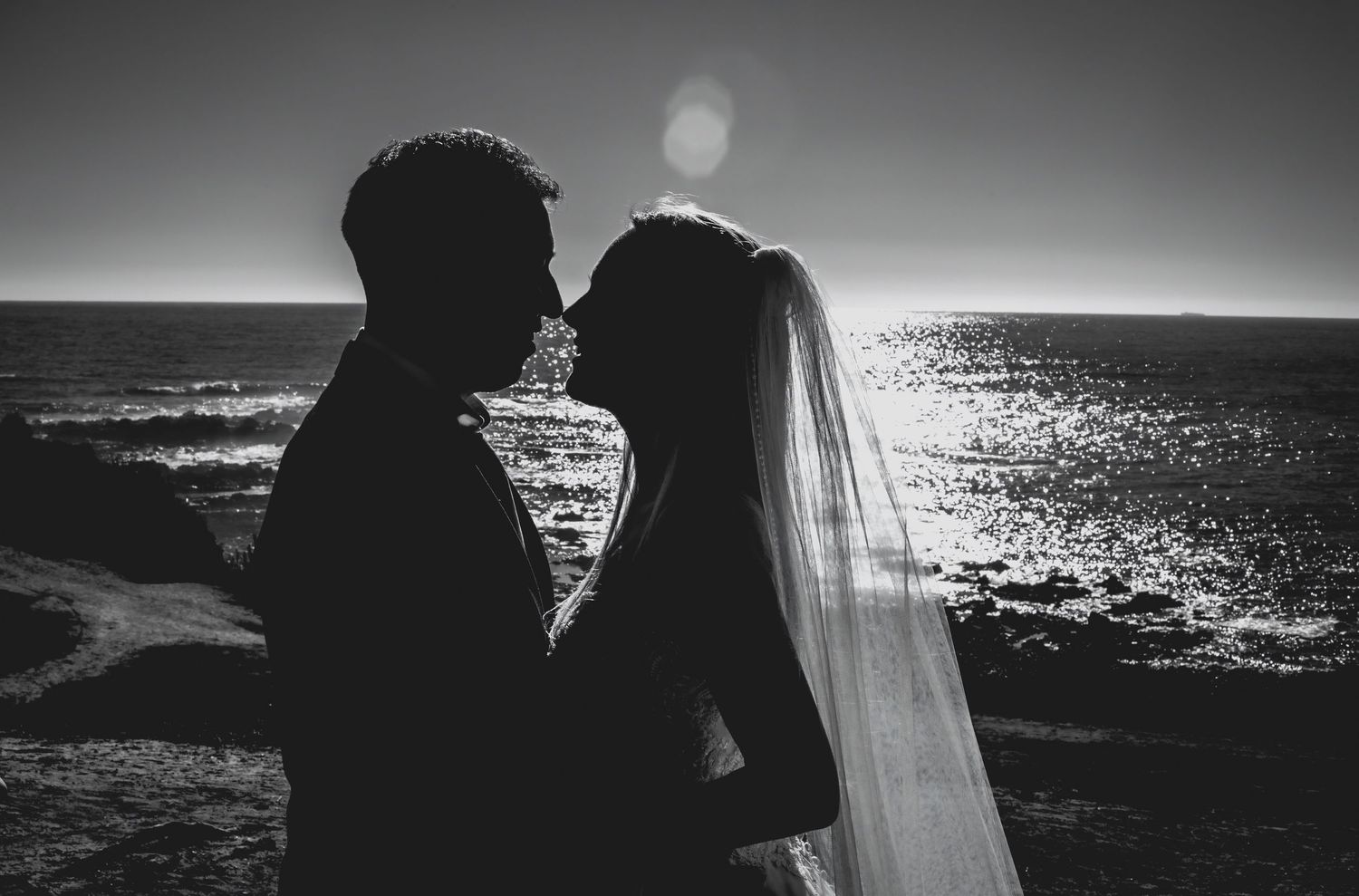 Romantic black and white silhouette of bride and groom nearly kissing with shimmering ocean in the background.