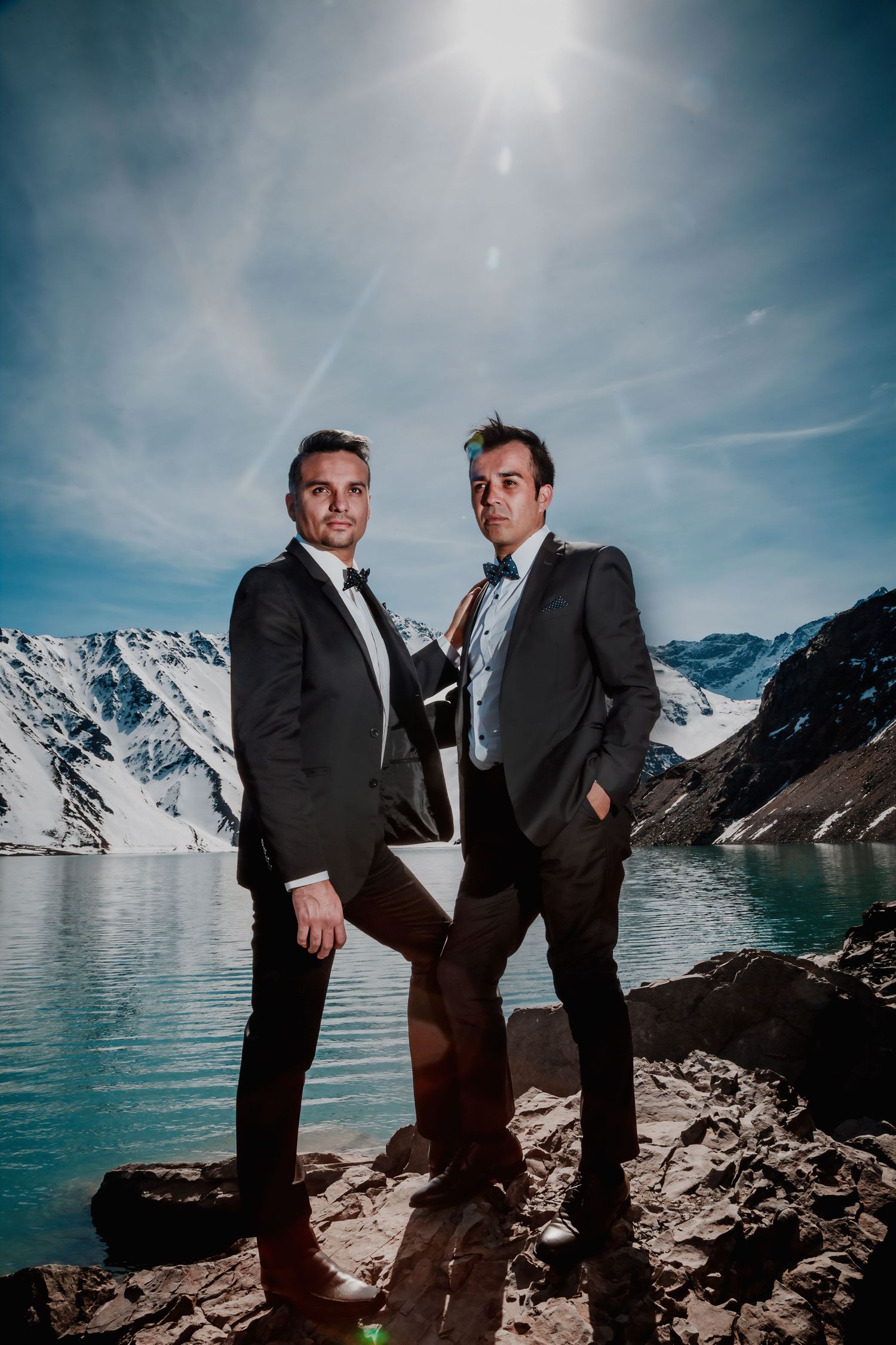 Two men in formal black tuxedos pose confidently by a mountain lake under a bright halo sun.