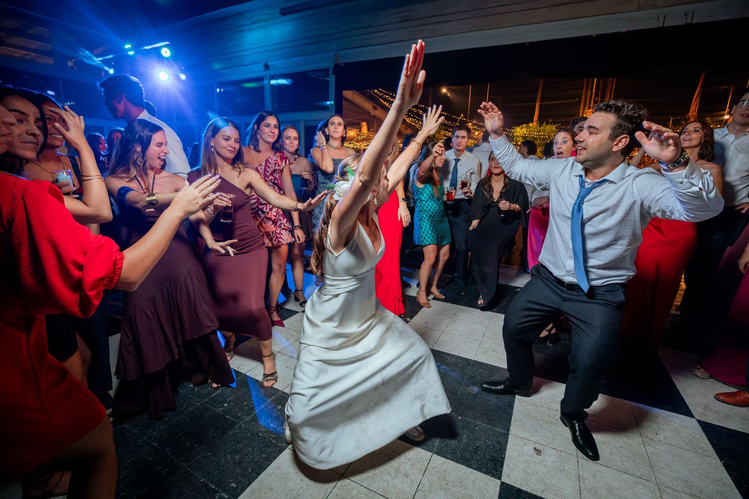 Bride in white gown dances energetically with groom and guests on illuminated wedding reception dance floor.