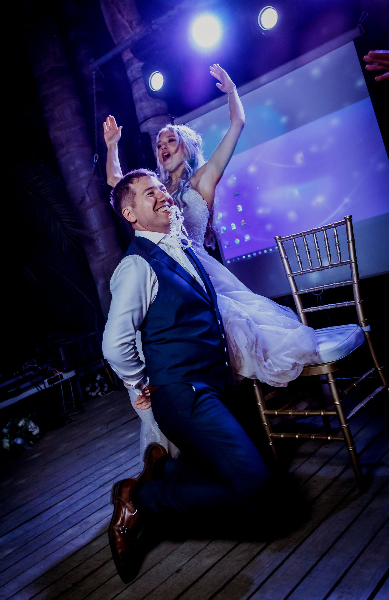 Groom sliding on knees while bride raises arms celebrating on stage at indoor wedding reception.