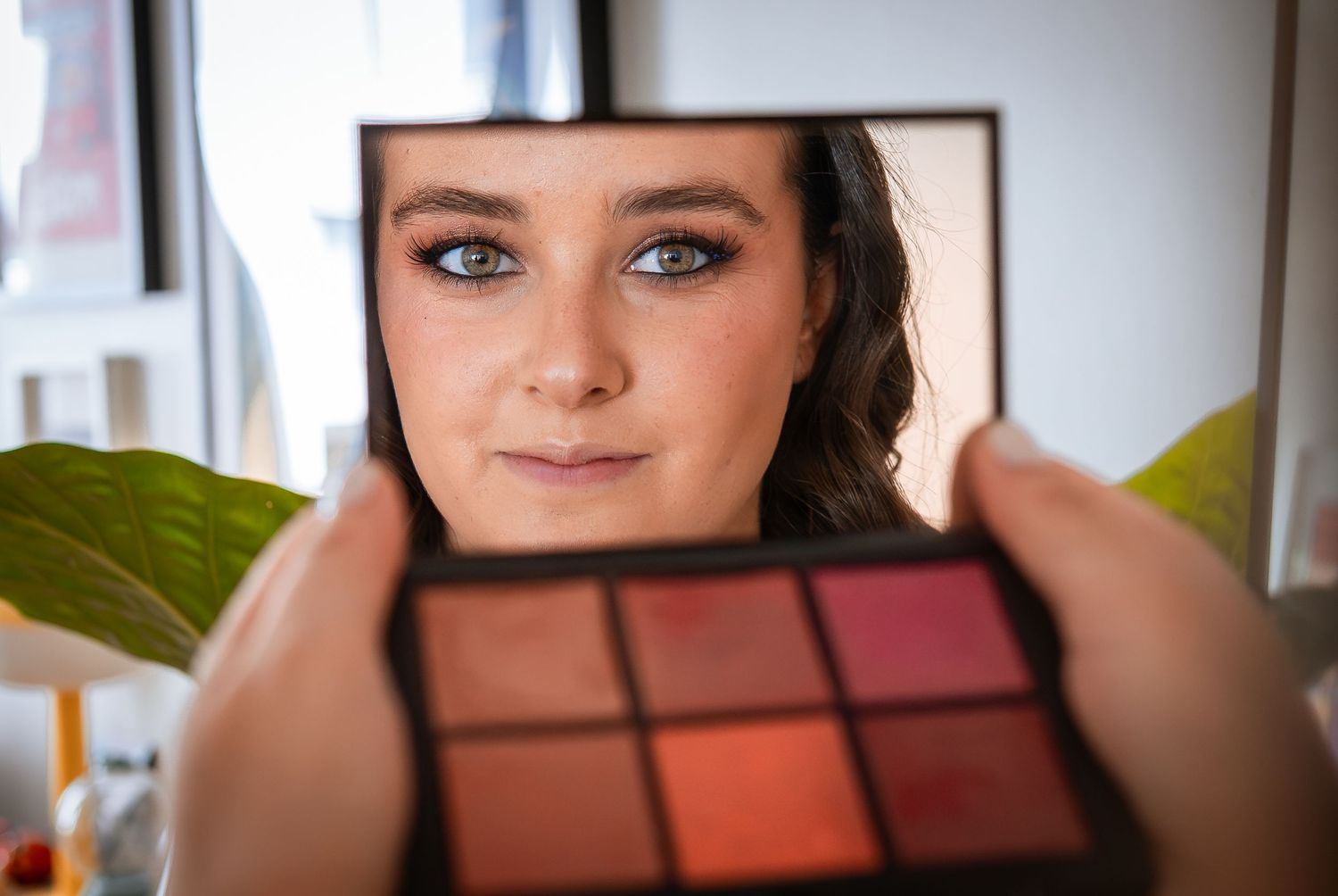 Young woman holds makeup palette in front of face reflecting in mirror indoors.