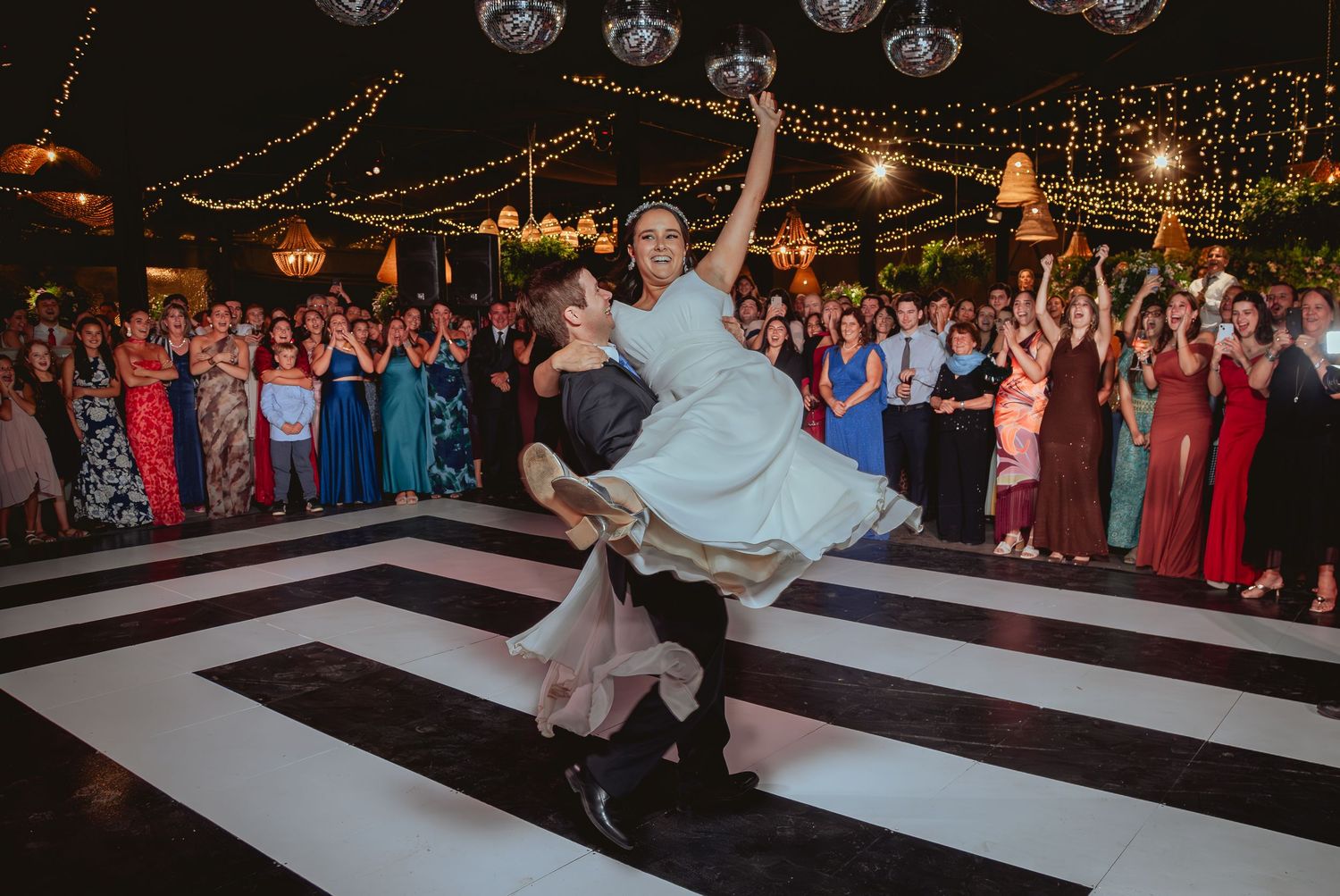 Groom lifts bride dramatically during energetic first dance at outdoor wedding reception with cheering guests.