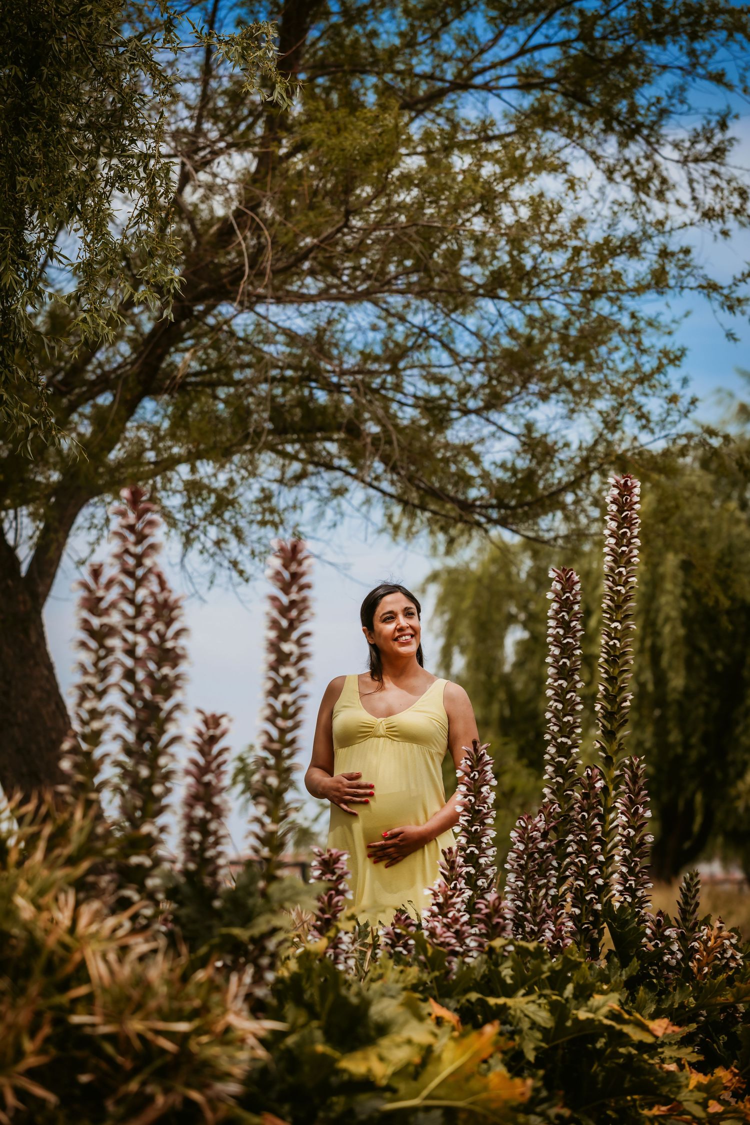 Smiling pregnant woman in yellow dress cradling baby bump among purple flowers and trees in a sunny outdoor garden.