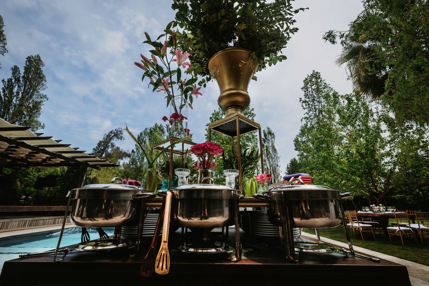 Elegant outdoor catering setup with gold urn centerpiece, chafing dishes, and lush garden backdrop near a pool.