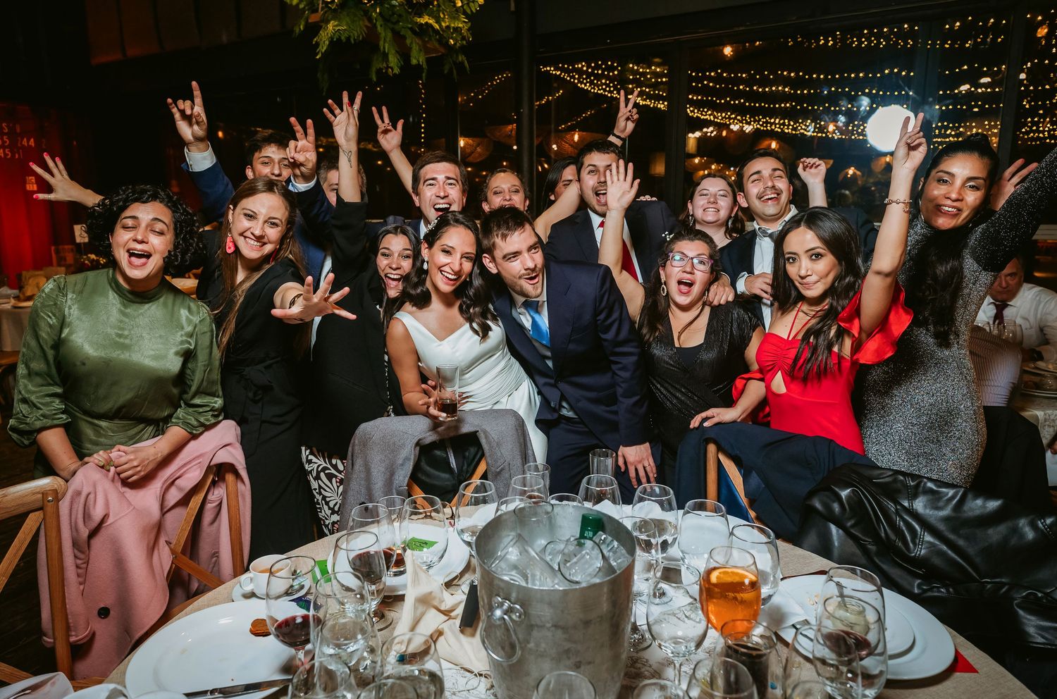 Joyful wedding reception group photo with bride and groom surrounded by celebrating guests at a festive dinner table.