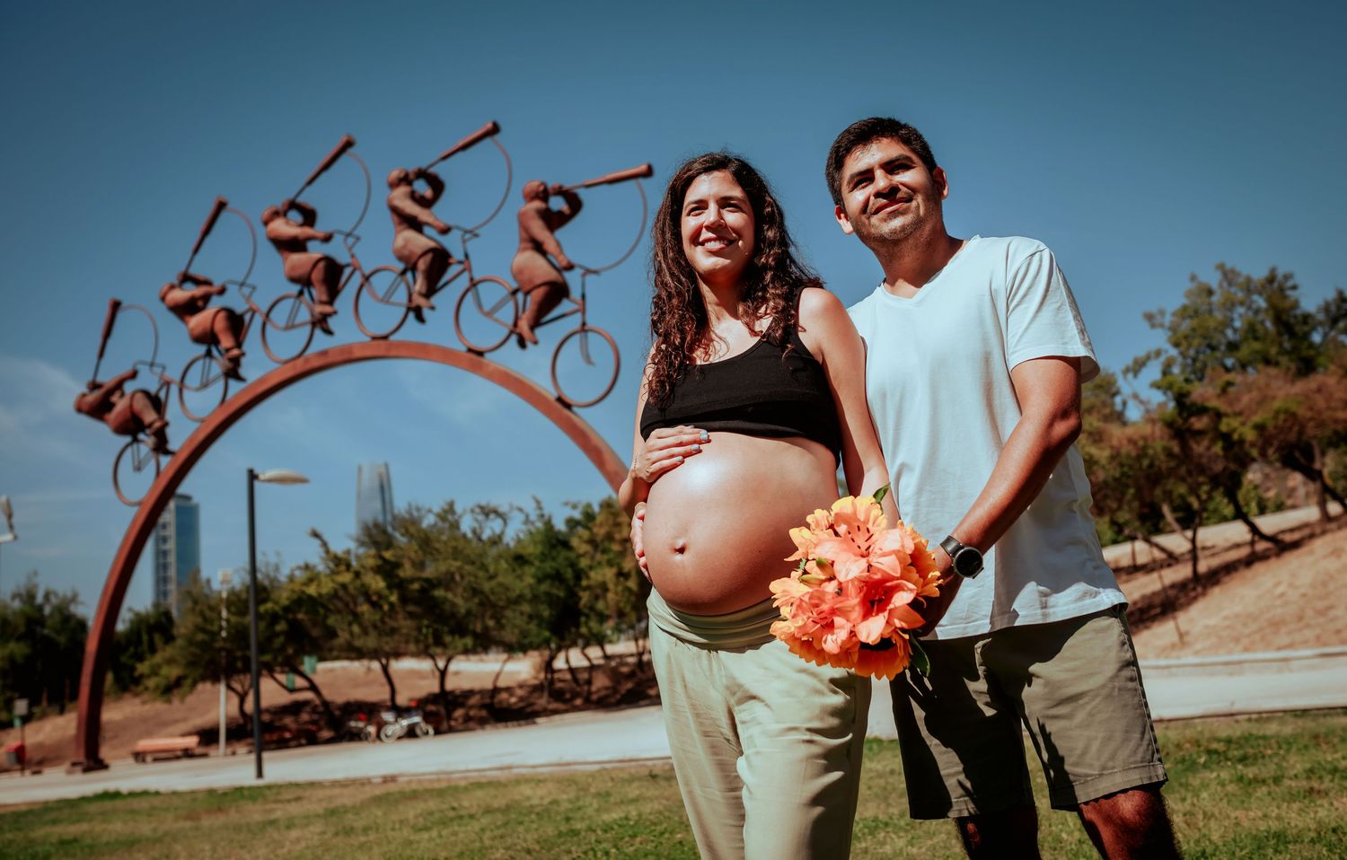 Pregnant couple standing in a park with a bicycle sculpture, woman holding orange flowers on a sunny day.