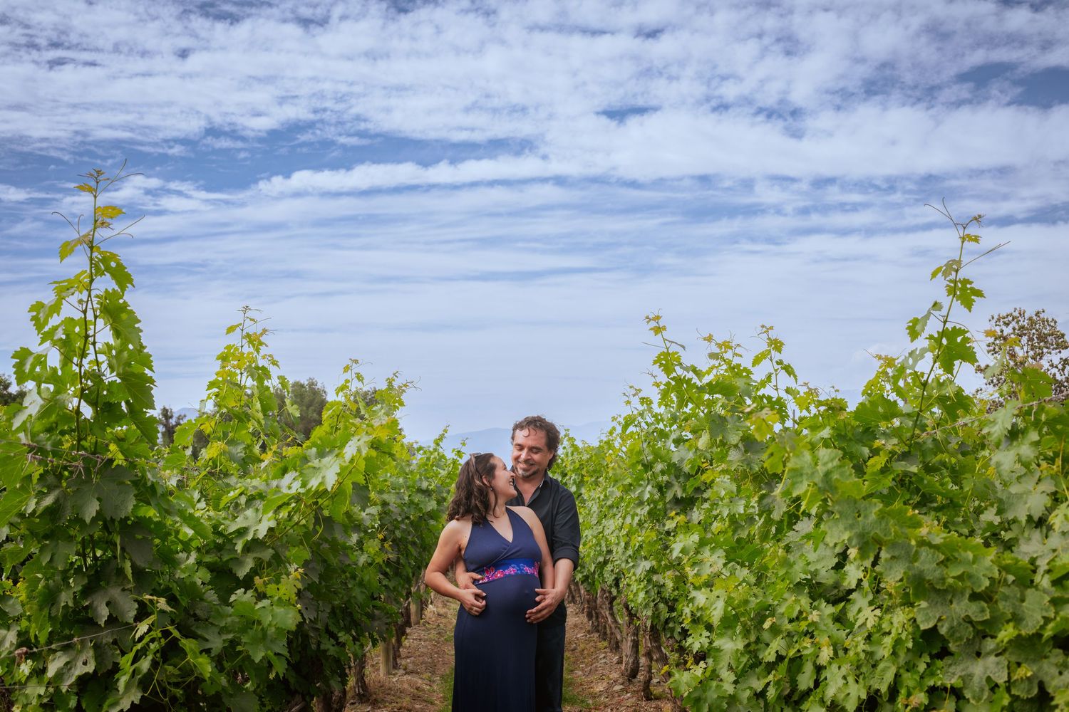 Couple embracing in lush green vineyard rows under blue sky with scattered clouds.