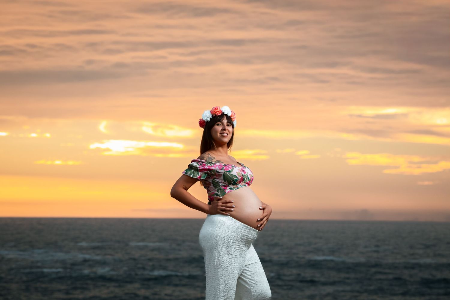 Smiling pregnant woman wearing flower crown stands by the sea during a golden sunset.