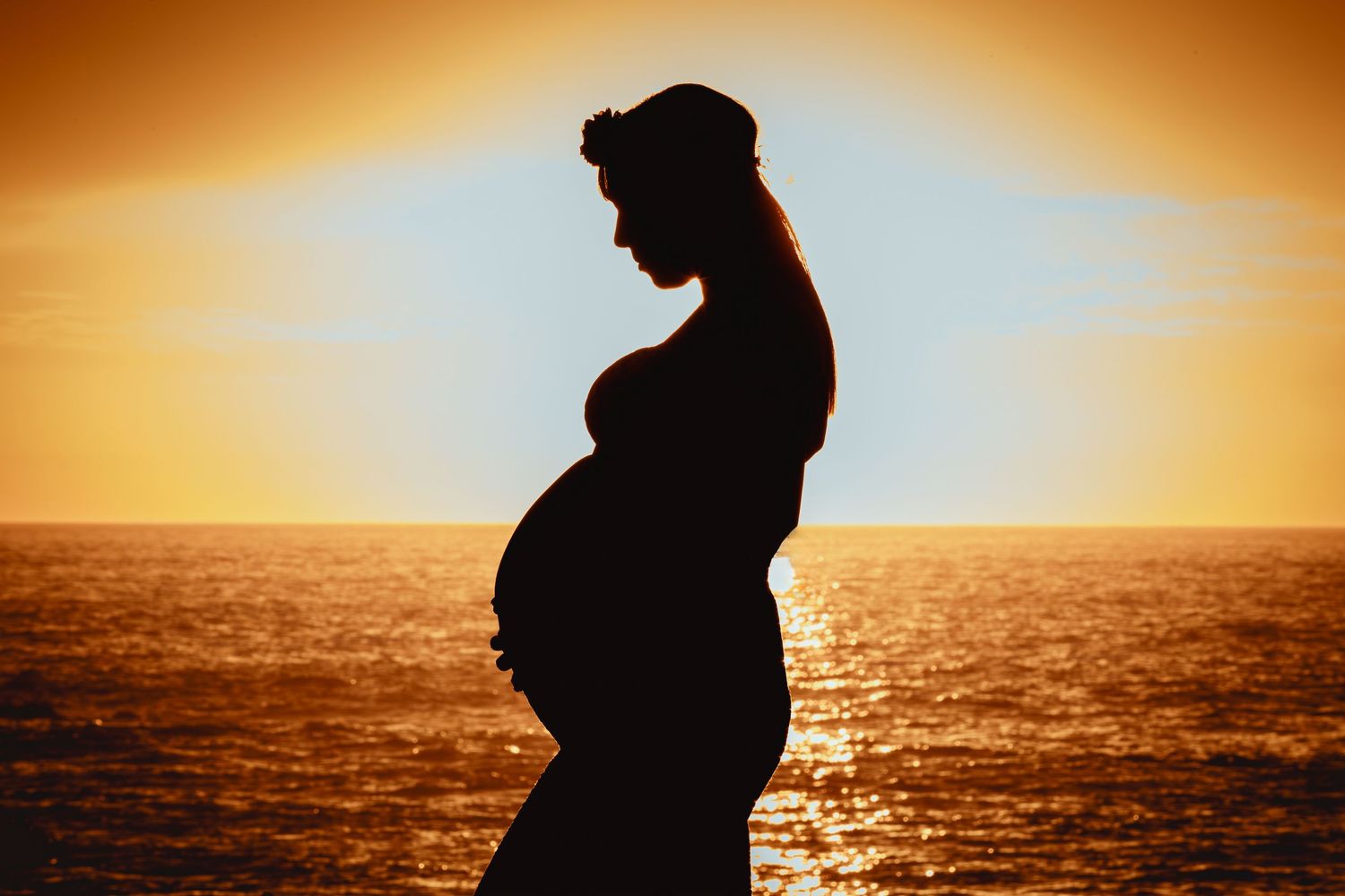 Pregnant woman silhouetted against a vibrant orange ocean sunset wearing a floral crown.