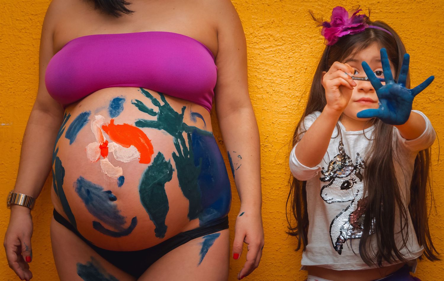 Pregnant woman stands beside a young girl showing off blue painted hands next to a colorfully painted baby bump.