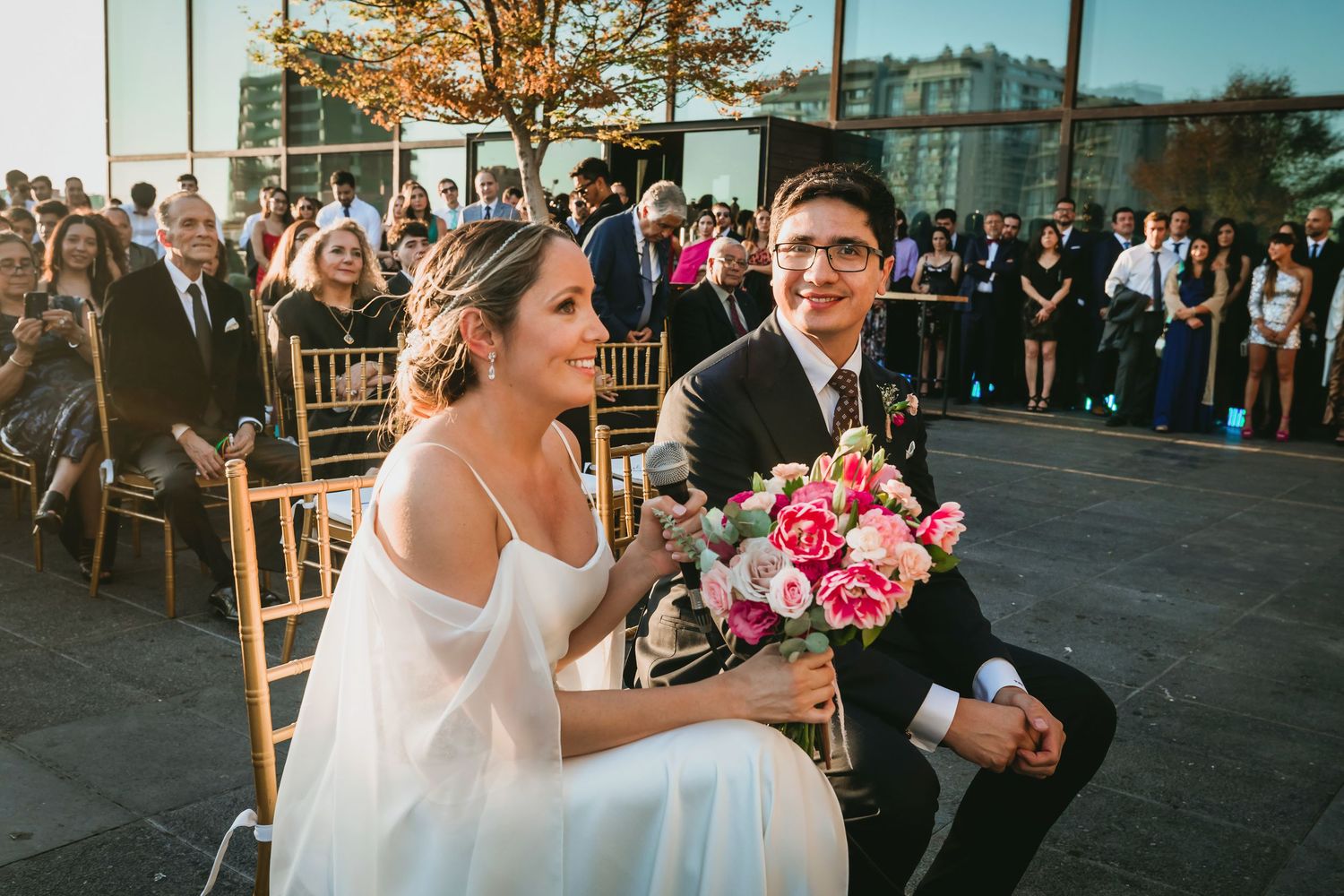 Smiling bride holds pink bouquet beside groom during sunny outdoor wedding ceremony.