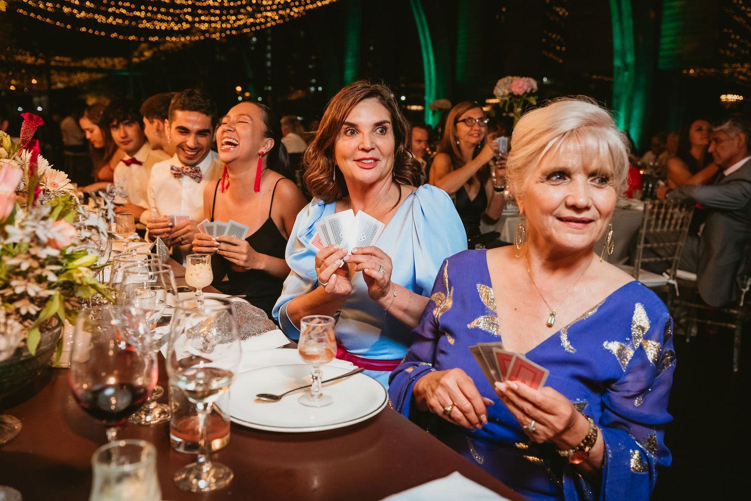 Wedding guests laughing and holding playing cards during a magic show at an outdoor reception.