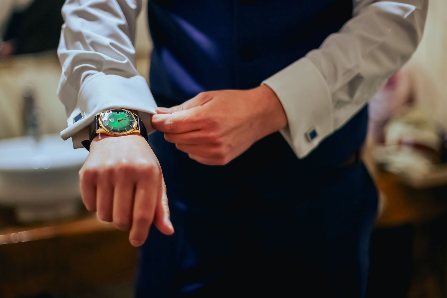 Groom adjusting gold watch with green face on his wrist wearing blue suit and white dress shirt.