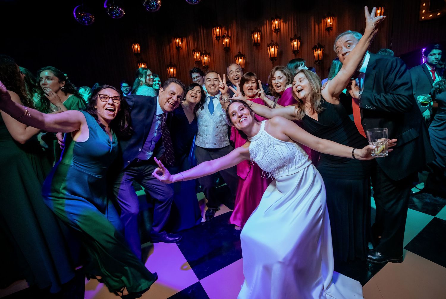 Bride in white gown poses with large group of celebrating wedding guests on colorful dance floor.