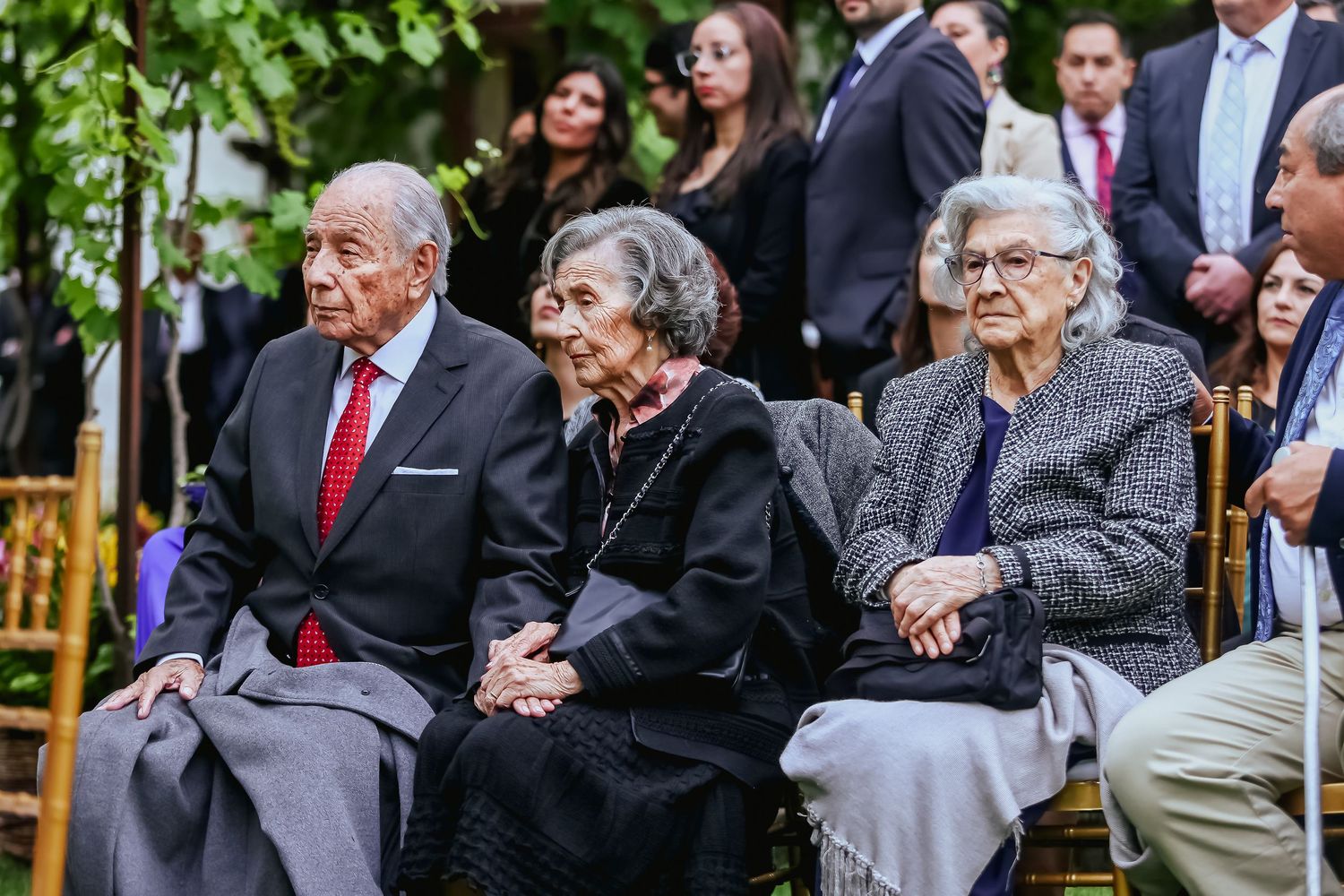 Three elderly wedding guests seated in gold chairs at outdoor ceremony, dressed in formal attire.