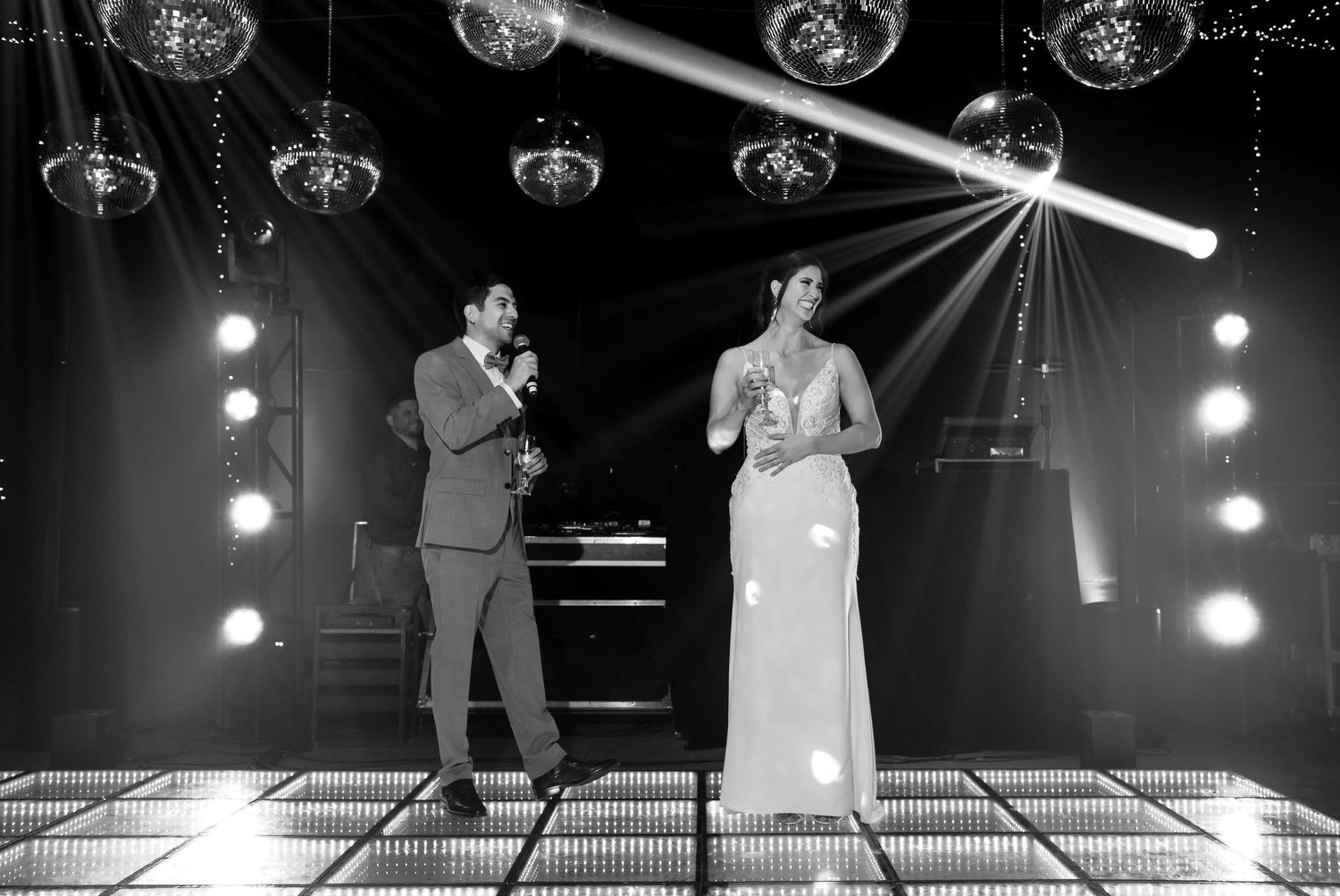 Black and white photo of a bride and groom sharing a microphone on a dance floor with dramatic stage lighting.