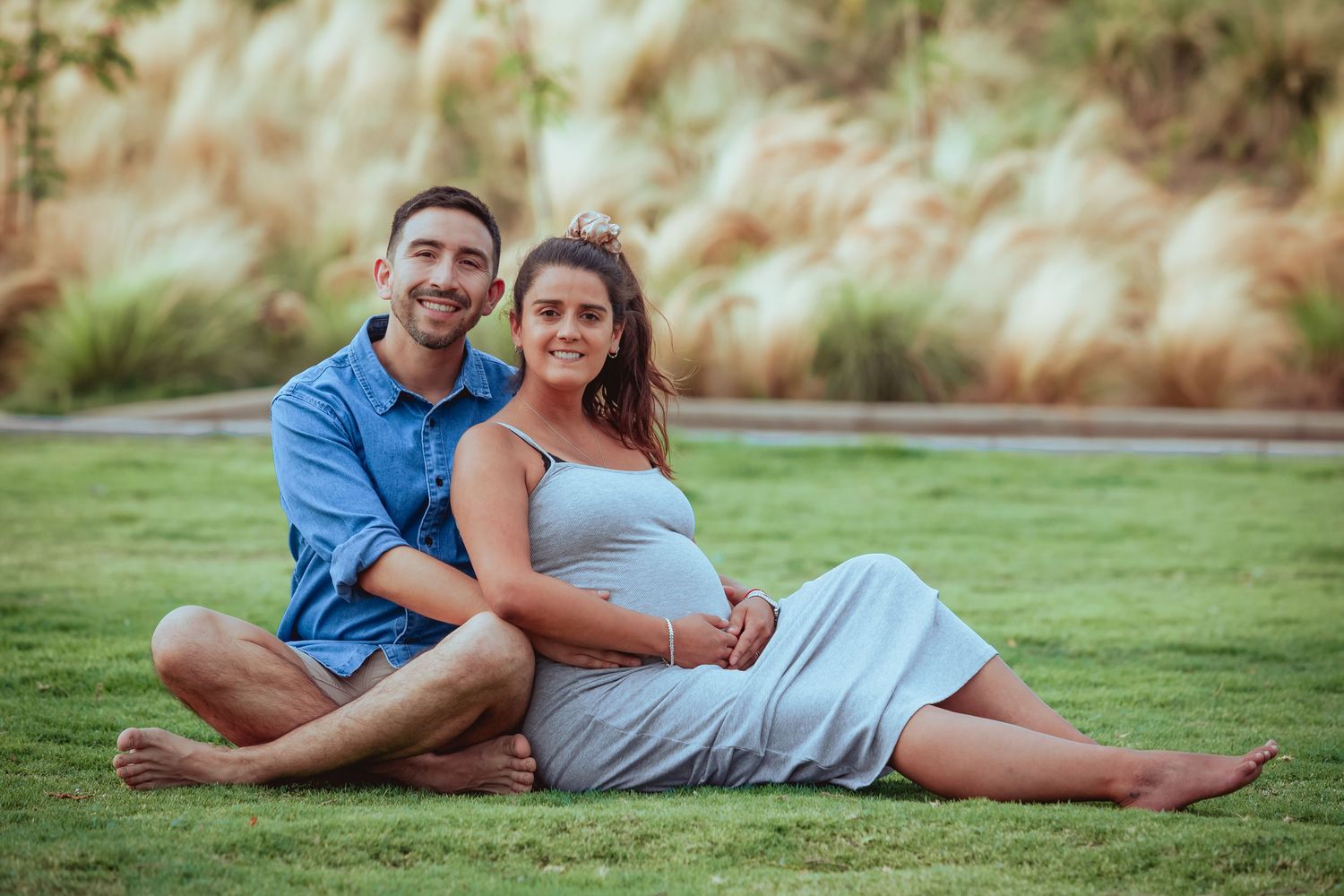 Couple sitting on grass, pregnant woman in grey dress leaning against partner in blue shirt, smiling.