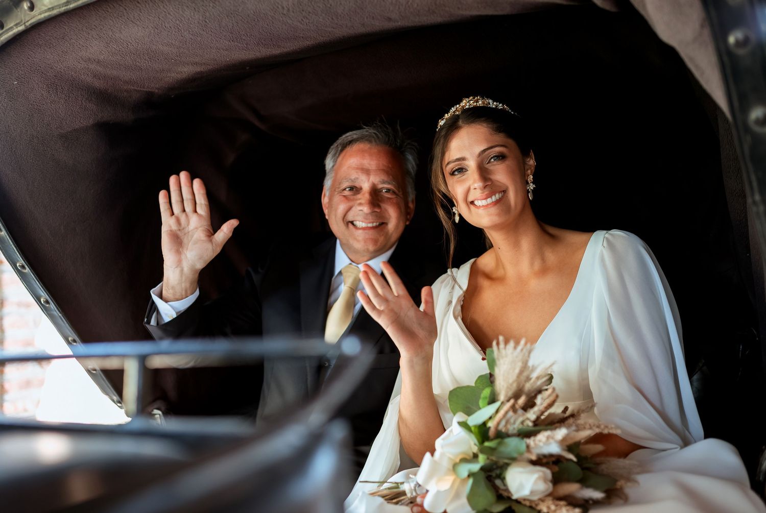 Bride and older man waving from a vintage carriage on wedding day.