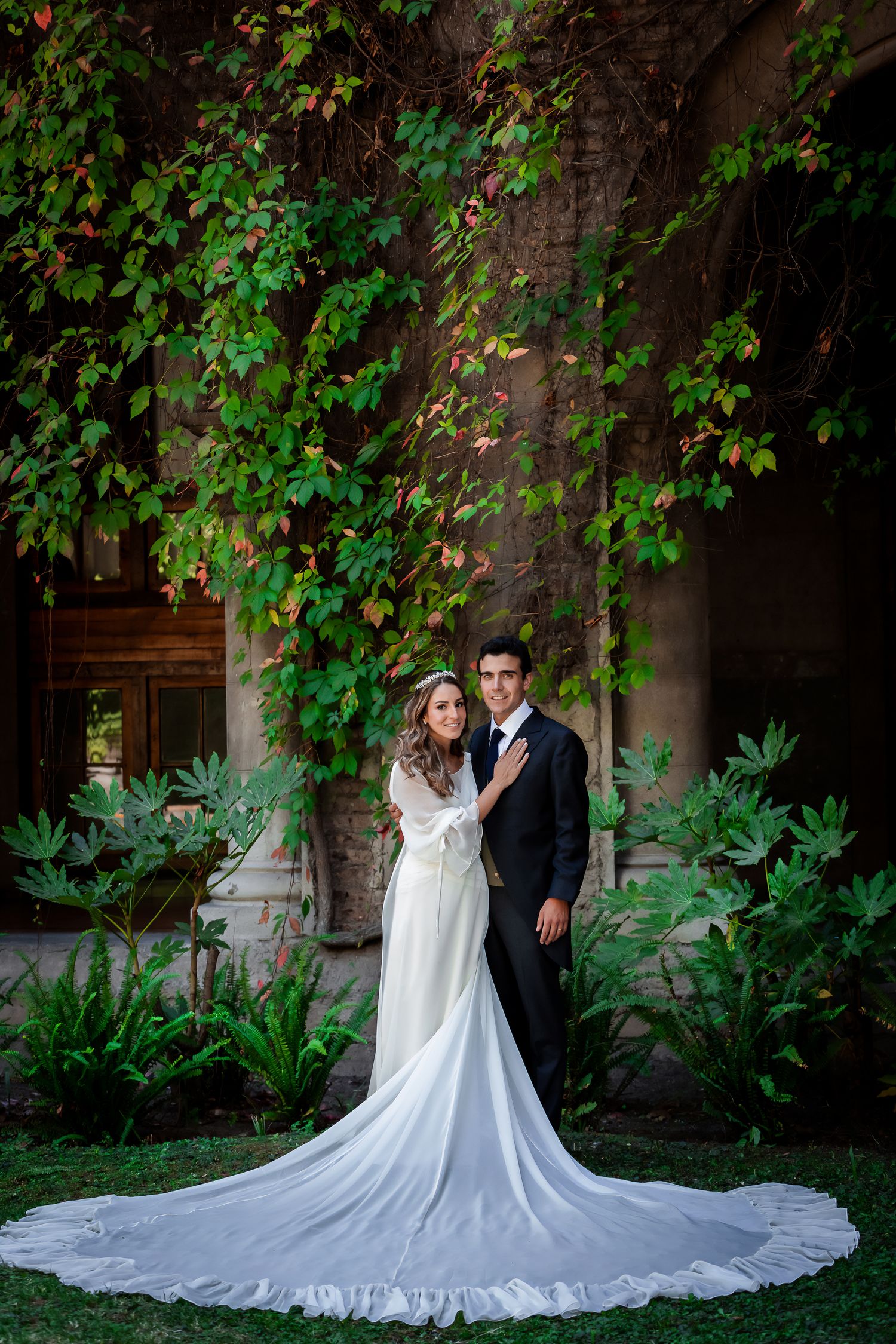 Bride and groom posing romantically before a lush green ivy-covered wall in a garden setting.