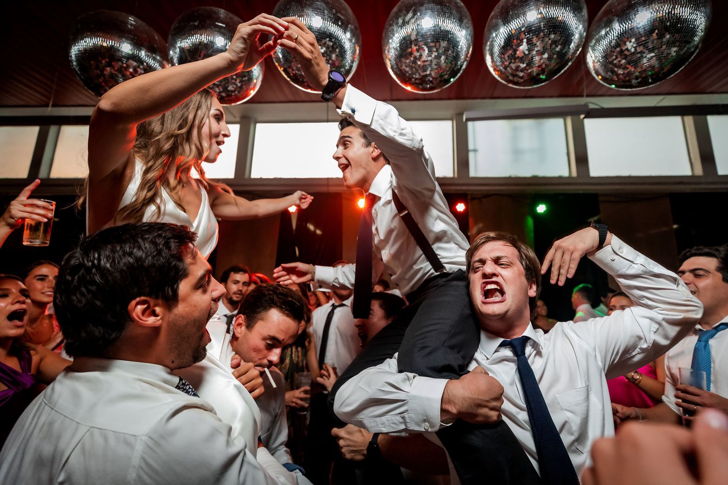 Bride and groom lifted by cheering guests on crowded dance floor under silver disco balls at wedding reception.