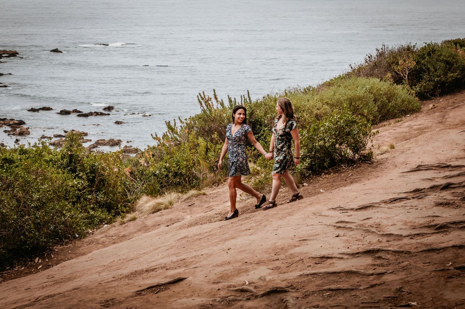 Two women in floral dresses walk hand in hand along a scenic coastal hillside path.