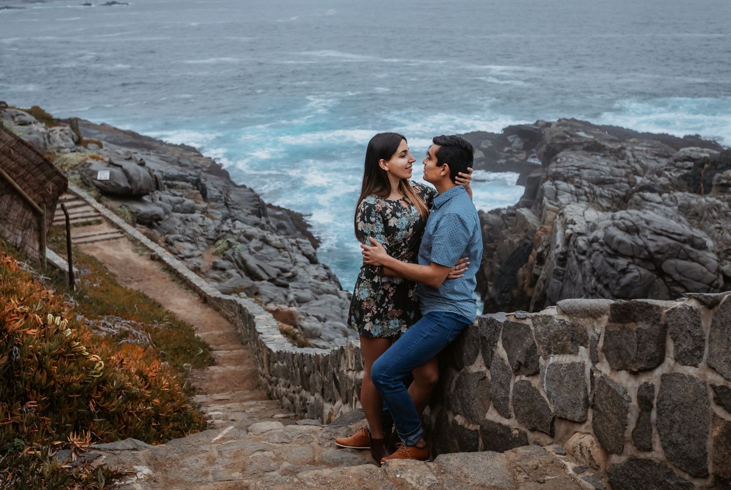 Romantic couple embracing near stone wall overlooking dramatic rocky coastline and blue ocean.