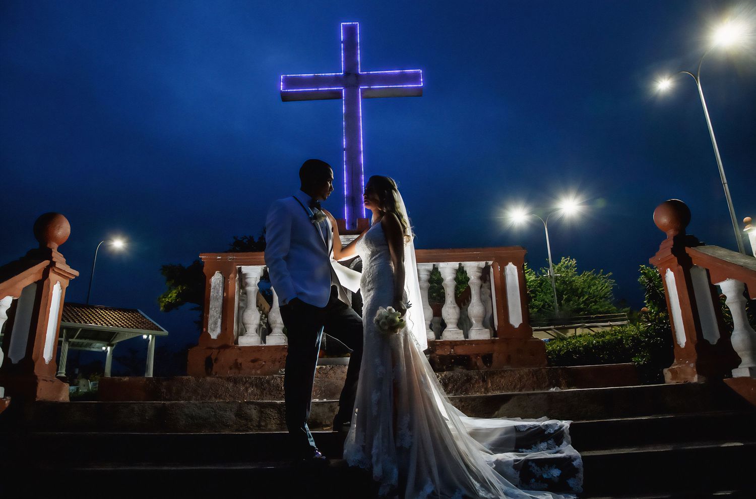 Bride and groom stand before an illuminated purple cross on church steps at night.
