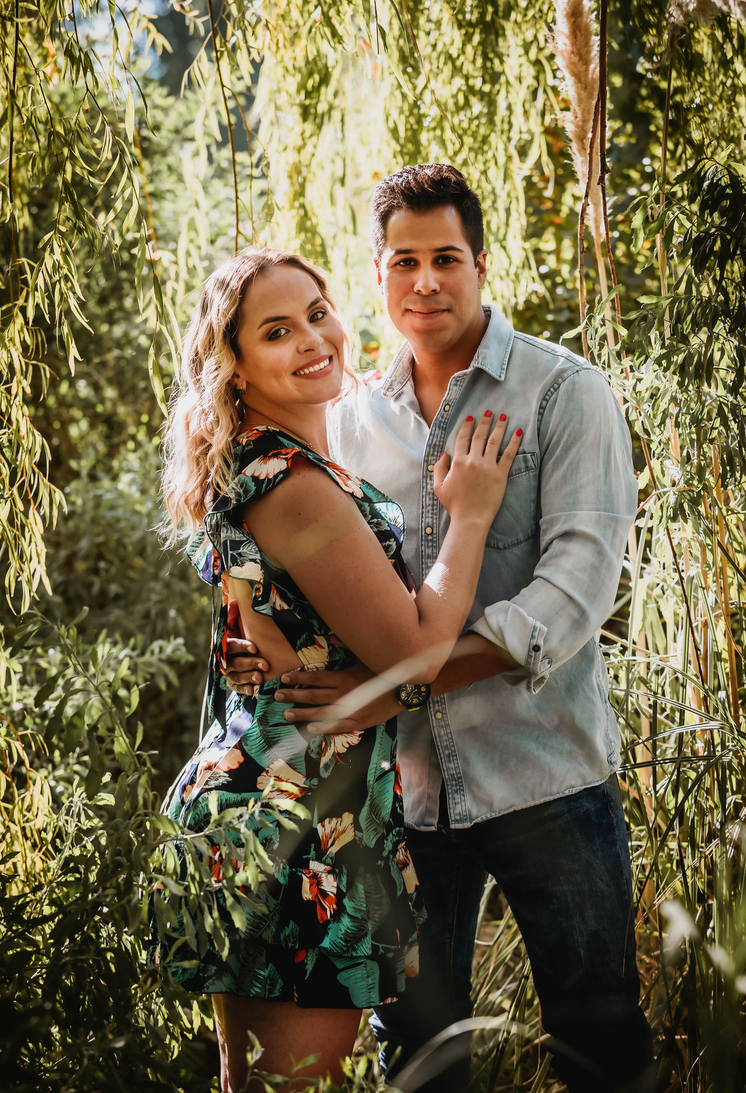 Smiling couple embracing outdoors under weeping willow tree, woman in floral dress, man in light blue shirt and jeans.
