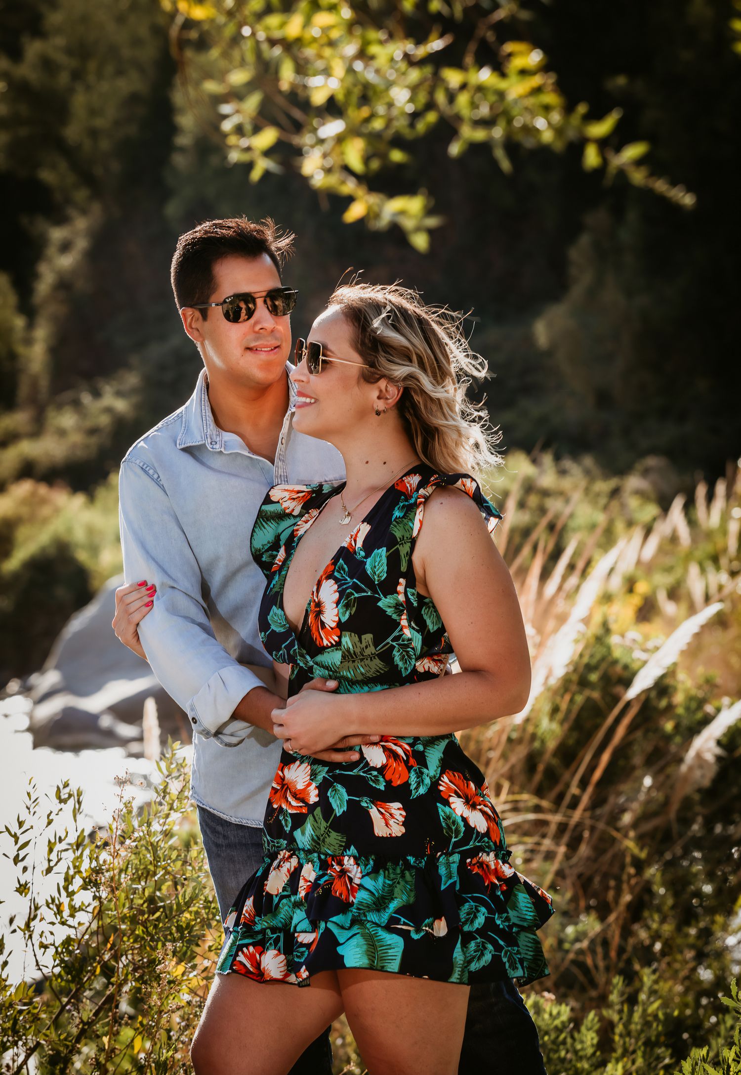 A couple in sunglasses embraces outdoors, woman in floral dress, man in light blue shirt, with lush greenery behind them.